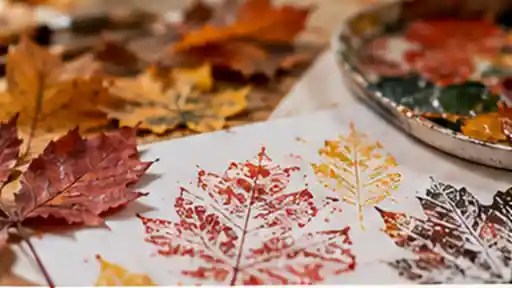 Autumn leaf prints in red, yellow and green on white paper beside fallen leaves and a tray of printing ink